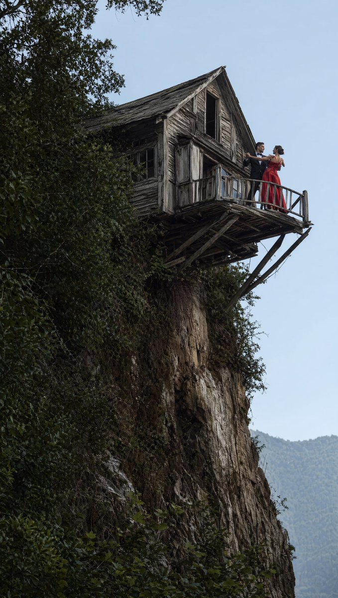 A dramatic vertical cinematic photograph of a weathered wooden house jutting out from the edge of a steep cliff, with a…