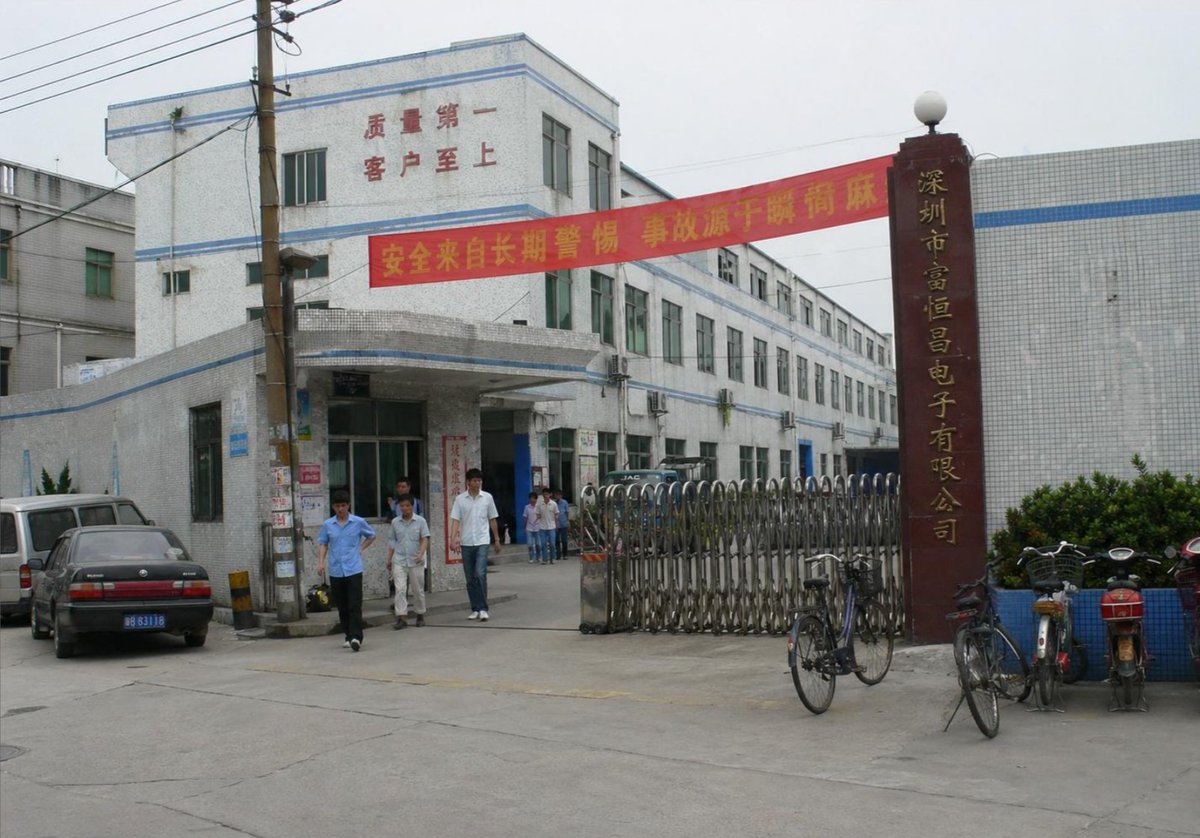 Create a photorealistic documentary-style street photograph of the entrance to a Chinese electronics factory in Shenzhe…