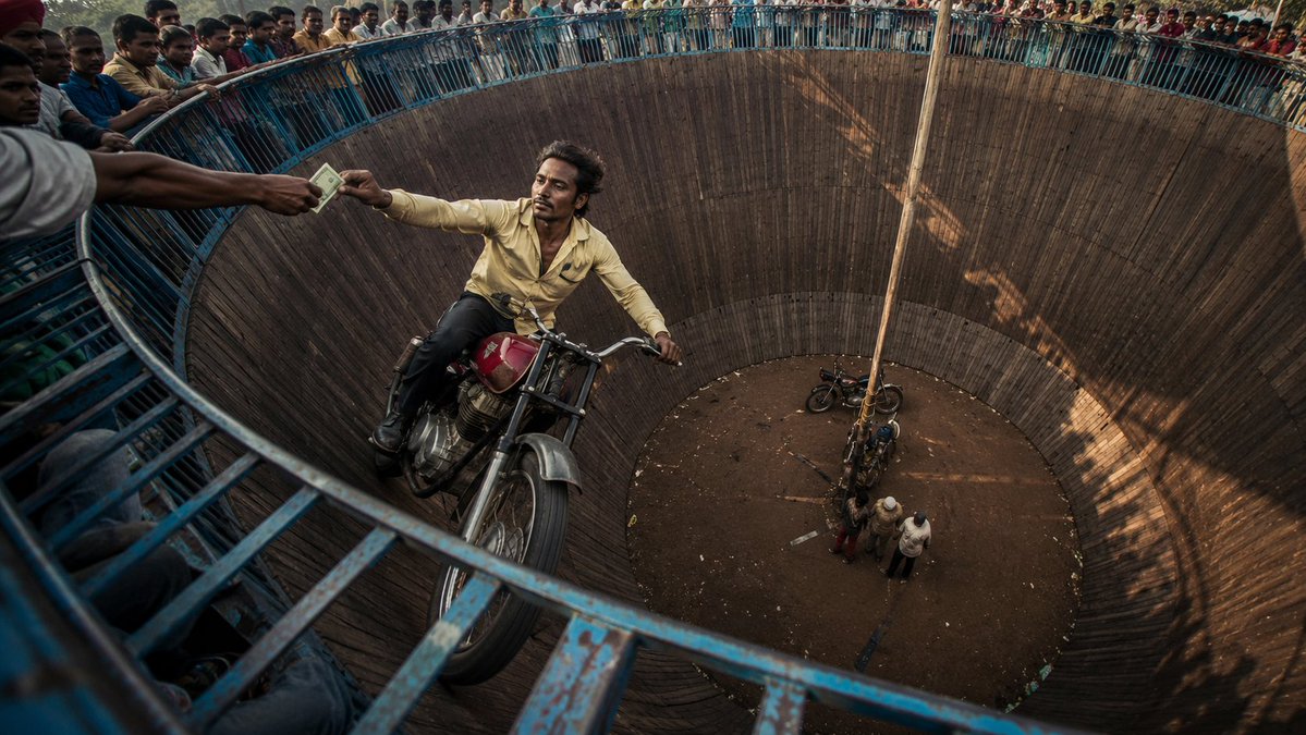 A cinematic wide-angle documentary photograph of a daring motorcycle stunt rider performing inside a massive circular W…
