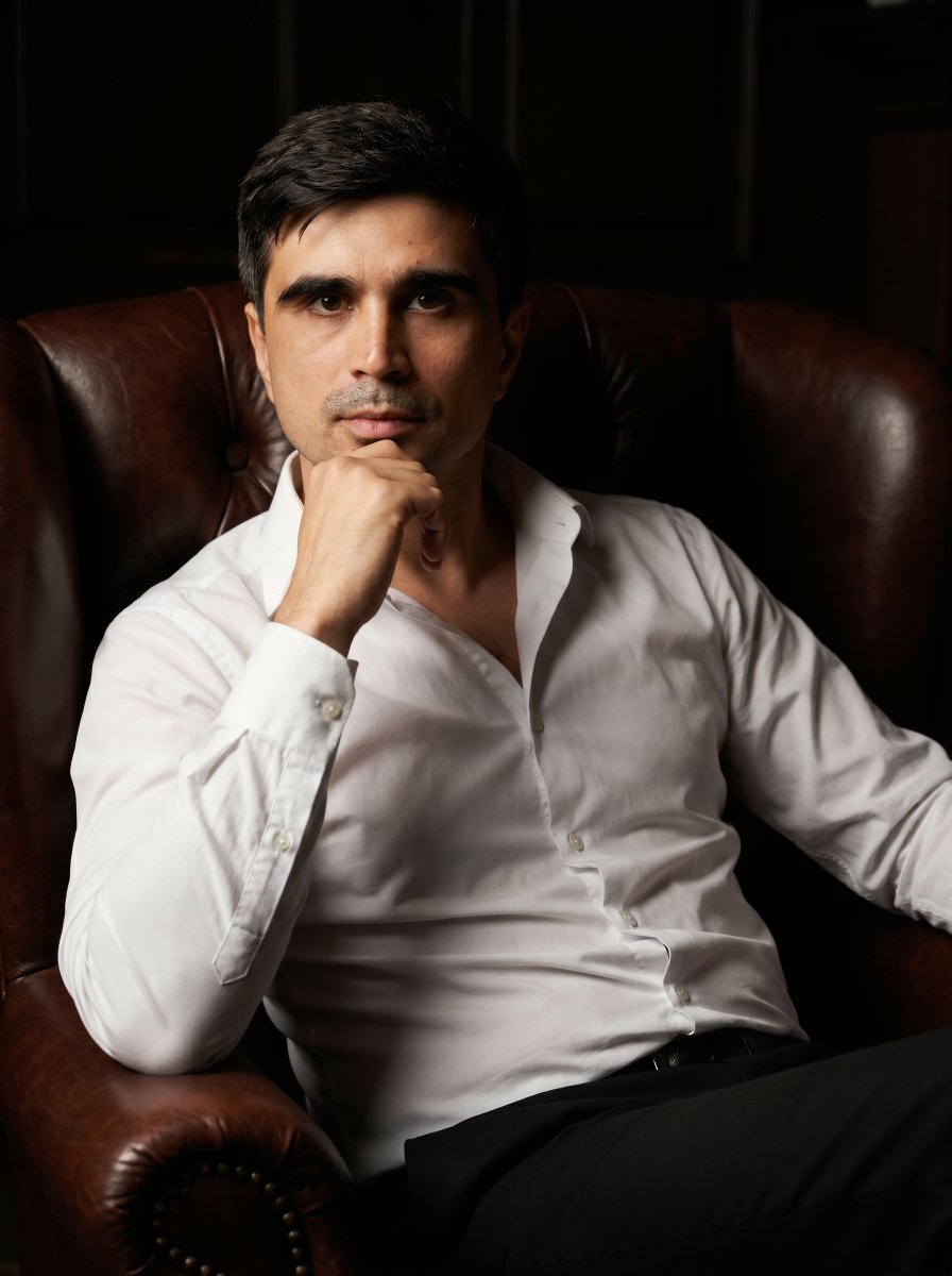 A cinematic portrait photo of a man sitting confidently in a dark, moody setting. He is dressed in a {argument name="cl…